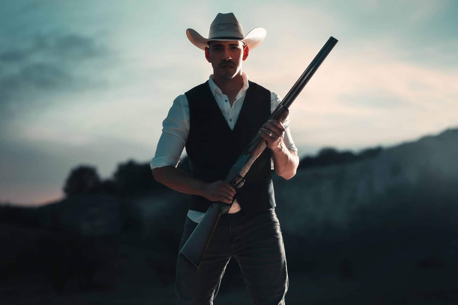 Homem com chapéu e rifle em cenário ao pôr do sol.
